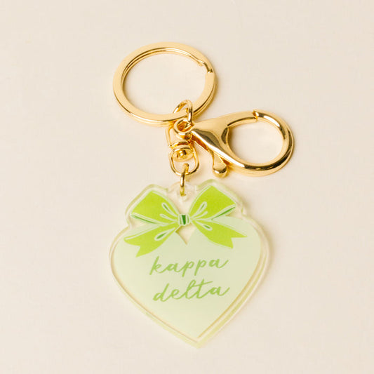 The Darling Effect's Kappa Delta Sweetheart Bow Acrylic Keychain features a gold heart, green bow, and "kappa delta" in light green script—a perfect sisterhood gift for any sorority member.