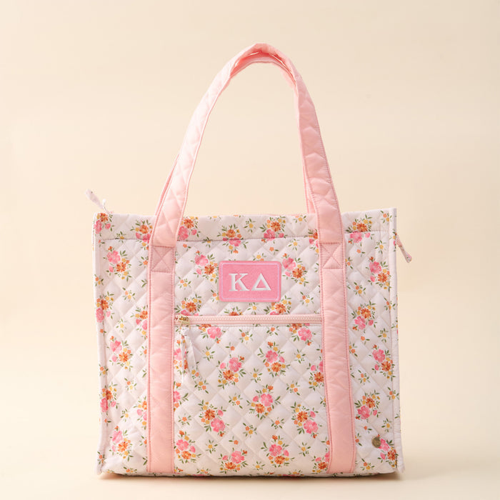 The Darling Effect’s Kappa Delta The Everyday Quilted Tote Bag features a floral design, light pink handles and trim, a front pocket with a pink ΚΔ patch, and a plain beige background—perfect for daily sorority style.