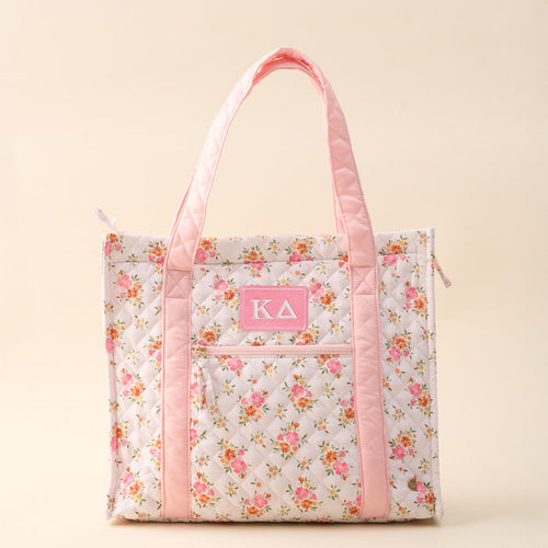 The Darling Effect’s Kappa Delta The Everyday Quilted Tote Bag features a floral design, light pink handles and trim, a front pocket with a pink ΚΔ patch, and a plain beige background—perfect for daily sorority style.