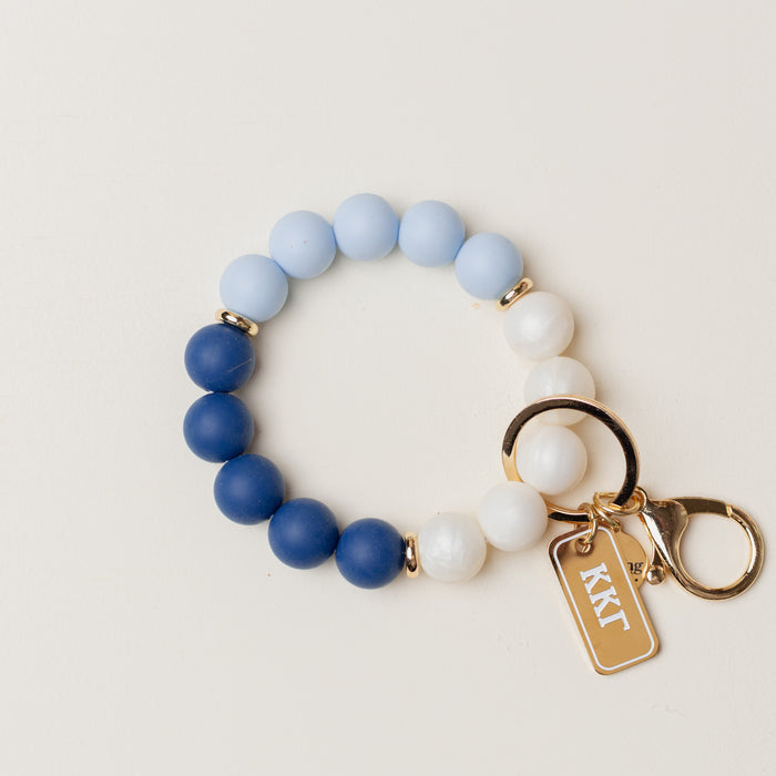 The Darling Effect Kappa Kappa Gamma Hands-Free Keychain Wristlet features alternating blue, light blue, and white silicone beads with gold accents, a gold keyring, and a rectangular gold tag engraved with KKΓ.