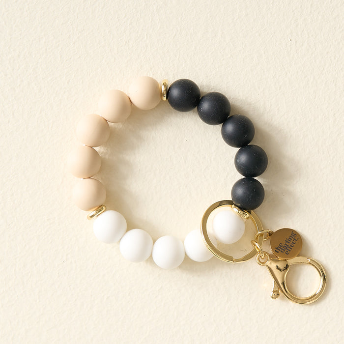 The Darling Effect’s Keep It Classy Hands-Free Keychain Wristlet features white, beige, and black silicone beads, gold hardware, a key ring, lobster clasp, and a “choose happy” charm—perfect for stylishly keeping your keys hands-free.