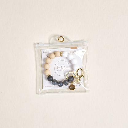 The Darling Effect Keep It Classy Hands-Free Keychain Wristlet features beige, white, and gray silicone beads with gold accents, a keyring, and comes in a clear zippered pouch for easy, stylish access.