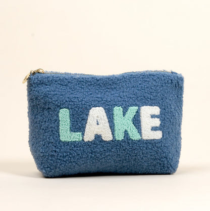 The Lake Teddy Pouch by the-darling-effect is a blue textured sherpa pouch featuring "LAKE" in large white and light green letters on the front. This cozy accessory stands upright against a plain off-white background.