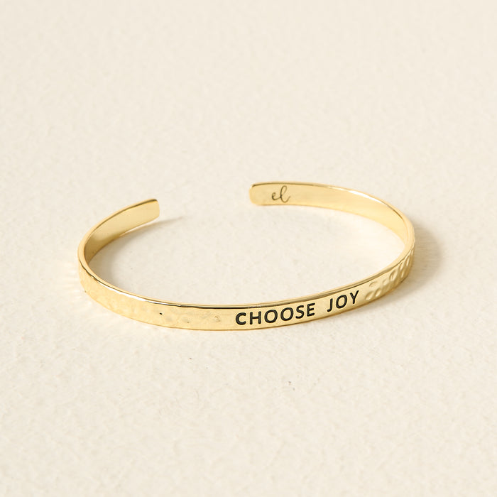 The Darling Effect's Little Reminder Cuff is a 14k gold, hammered adjustable bracelet engraved with CHOOSE JOY in black letters, photographed on a light textured surface.