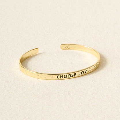The Darling Effect's Little Reminder Cuff is a 14k gold, hammered adjustable bracelet engraved with CHOOSE JOY in black letters, photographed on a light textured surface.