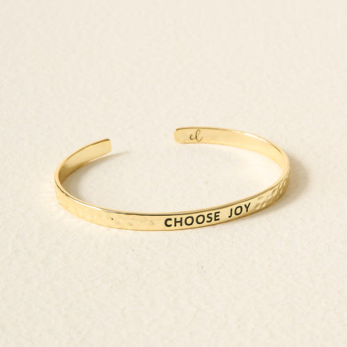 The Darling Effect's Little Reminder Cuff is a 14k gold, hammered adjustable bracelet engraved with CHOOSE JOY in black letters, photographed on a light textured surface.
