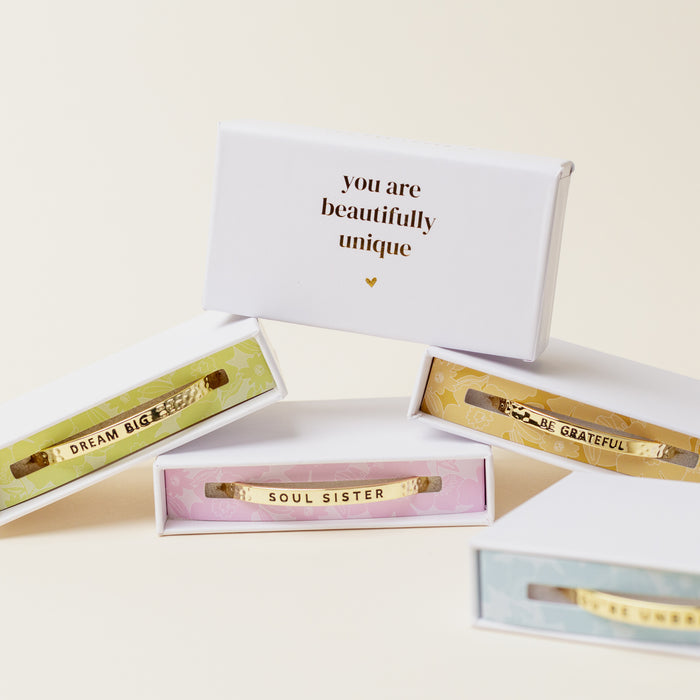 Four adjustable Little Reminder Cuff bracelets by The Darling Effect, in colorful boxes engraved with DREAM BIG, SOUL SISTER, BE GRATEFUL. A white box behind them reads "you are beautifully unique" with a small gold heart beneath.