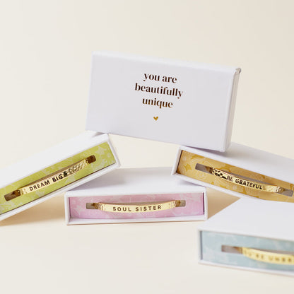 Four adjustable Little Reminder Cuff bracelets by The Darling Effect, in colorful boxes engraved with DREAM BIG, SOUL SISTER, BE GRATEFUL. A white box behind them reads "you are beautifully unique" with a small gold heart beneath.