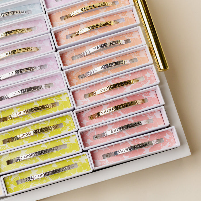A display tray showcases The Darling Effect's Little Reminder Cuff alongside decorative boxes of hair clips, each engraved with motivational phrases like SHINE BRIGHT, CHOOSE JOY, MAMA, and BESTIE.