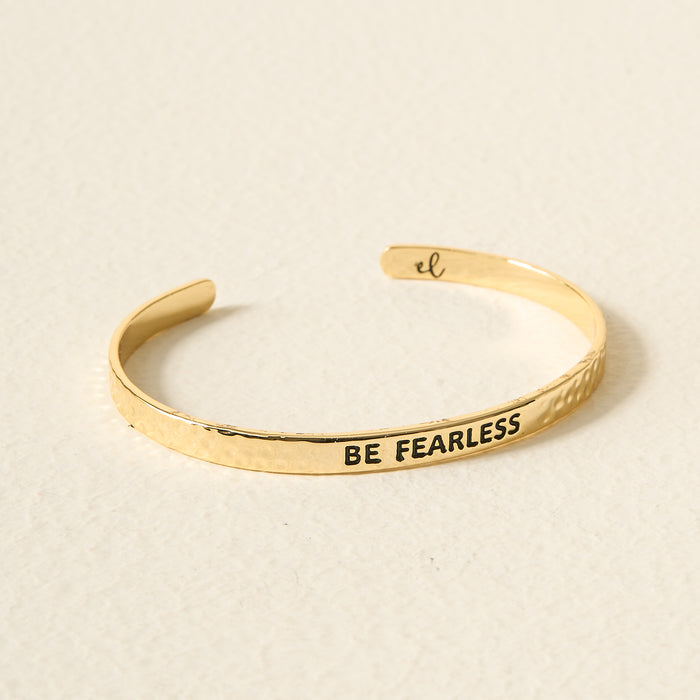 The Darling Effect Little Reminder Cuff is a 14k gold adjustable bracelet engraved with BE FEARLESS on the outside and a small “el” inside, shown on a light surface.