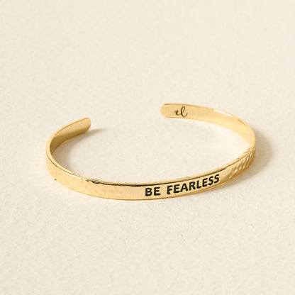 The Darling Effect Little Reminder Cuff is a 14k gold adjustable bracelet engraved with BE FEARLESS on the outside and a small “el” inside, shown on a light surface.