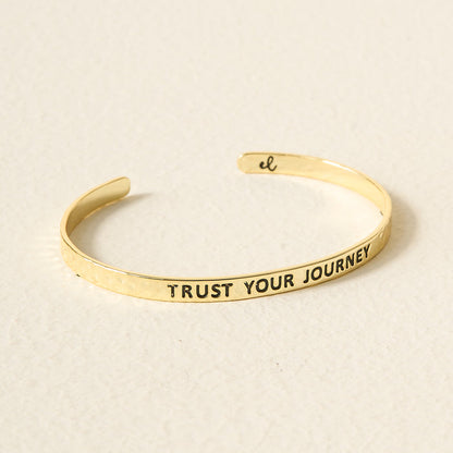 The Darling Effect's Little Reminder Cuff is a 14k gold, hammered, open-ended bracelet engraved with TRUST YOUR JOURNEY in black letters. This adjustable mantra cuff rests on a light, textured surface.