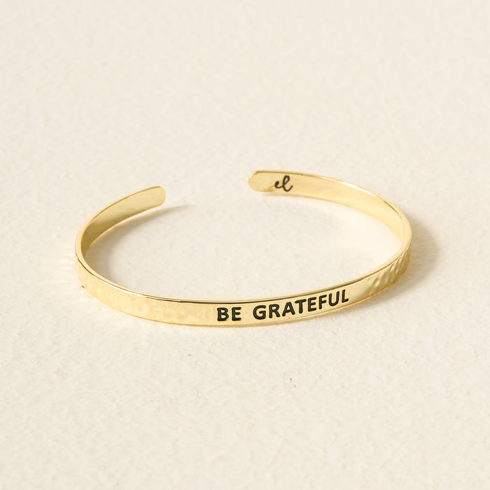 The Darling Effect's Little Reminder Cuff is a 14k gold adjustable bracelet engraved with BE GRATEFUL outside and a script "el" inside, displayed on a light-colored surface.