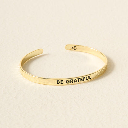 The Darling Effect's Little Reminder Cuff is a 14k gold adjustable bracelet engraved with BE GRATEFUL outside and a script "el" inside, displayed on a light-colored surface.