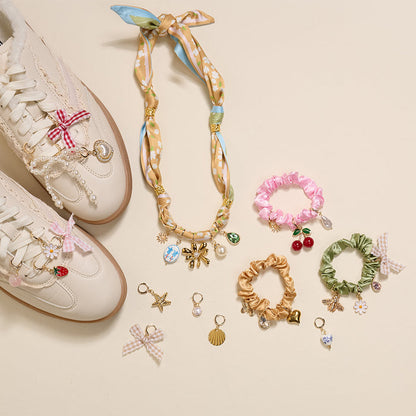 A flat lay on beige features white sneakers with charms, a gold and blue statement necklace, four colorful scrunchie charm sets, and The Darling Effect's Midnight Moments The Ultimate Charm Pack with flower, shell, star, cherry, and bow pendants.