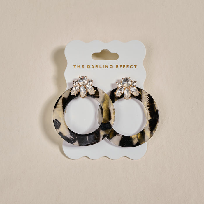 The Darling Effect Midnight Opal Glow Getter Earrings—circular, glossy acetate with crystal flower studs—are displayed on a white card labeled THE DARLING EFFECT against a neutral background.