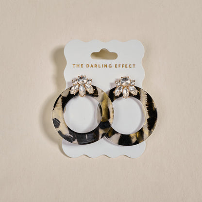 The Darling Effect Midnight Opal Glow Getter Earrings—circular, glossy acetate with crystal flower studs—are displayed on a white card labeled THE DARLING EFFECT against a neutral background.