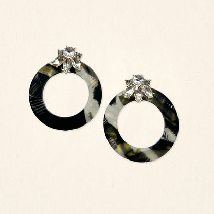 The Darling Effect's Midnight Opal Glow Getter Earrings—round, black and gray marbled with clear flower-shaped gems—are displayed on a light beige background.