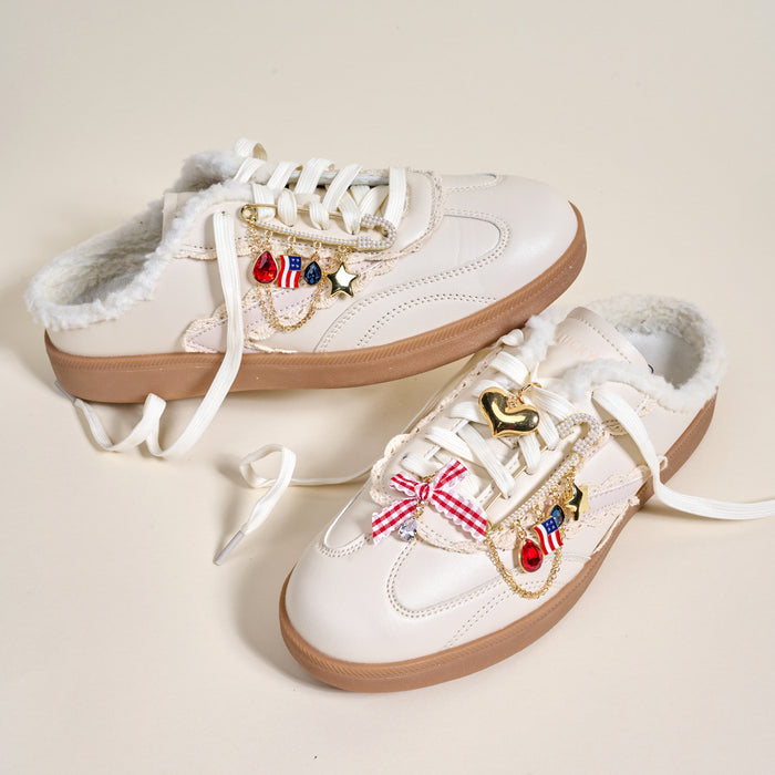 Two white slip-on sneakers with brown soles and fuzzy lining, styled with The Darling Effect's Miss Americana Dazzle Shoe Charm Pack—featuring a gold heart, stars, red bow, flag, and gem charms for a playful look.