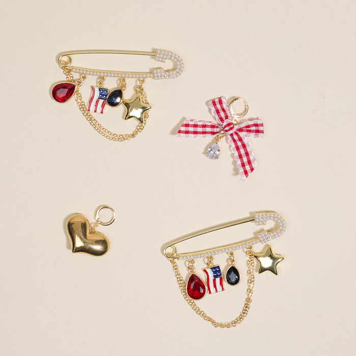 The Darling Effect’s Miss Americana Dazzle Shoe Charm Pack features three gold safety pin brooches with heart, star, flag, and shoe charms, plus colorful gems and bows—unique footwear accessories on a beige background.