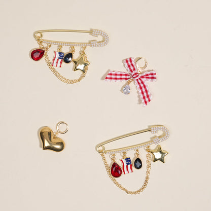 The Darling Effect’s Miss Americana Dazzle Shoe Charm Pack features three gold safety pin brooches with heart, star, flag, and shoe charms, plus colorful gems and bows—unique footwear accessories on a beige background.