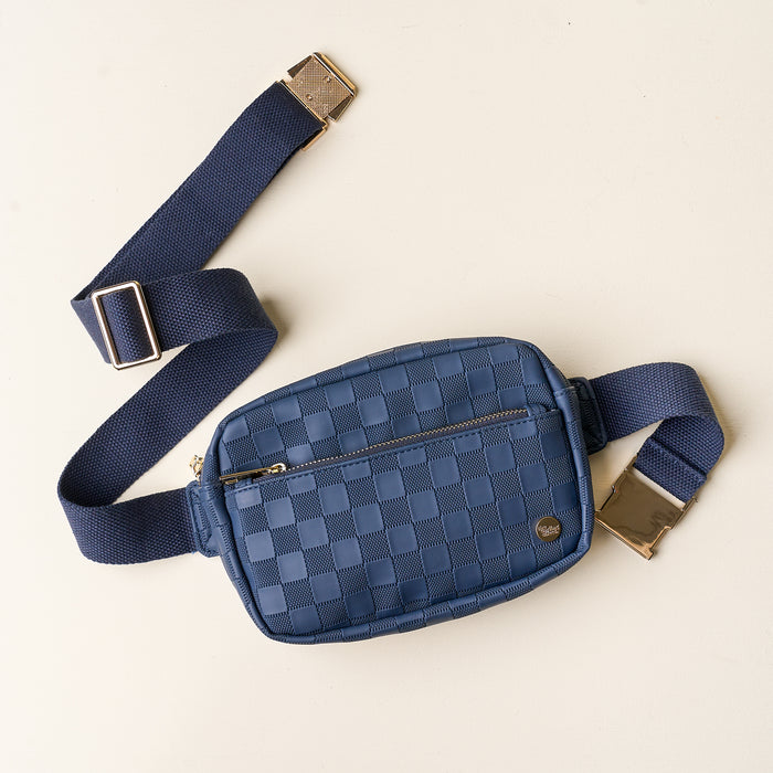 The Darling Effect’s Navy Urban Check Belt Bag features a blue checkered design, front zipper pocket, gold buckle on an adjustable navy strap, and is crafted from vegan leather, shown on a light beige surface.
