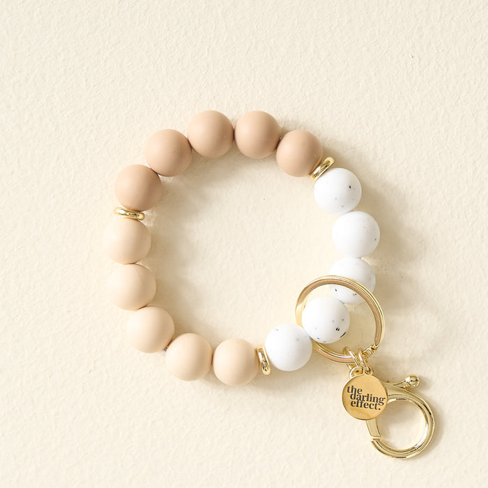 The Darling Effect Oatmilk Latte Hands-Free Keychain Wristlet features large beige and white silicone beads, gold spacer beads, a gold keyring, and an engraved charm for convenient, hands-free use. Displayed on a light cream surface.