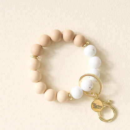 The Darling Effect Oatmilk Latte Hands-Free Keychain Wristlet features large beige and white silicone beads, gold spacer beads, a gold keyring, and an engraved charm for convenient, hands-free use. Displayed on a light cream surface.