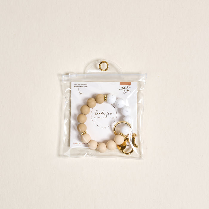 The Darling Effect Oatmilk Latte Hands-Free Keychain Wristlet features beige and white silicone beads with a gold clasp, packaged in a clear zippered pouch on a light beige background for stylish convenience.