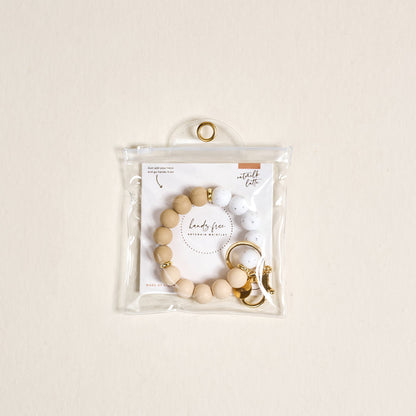 The Darling Effect Oatmilk Latte Hands-Free Keychain Wristlet features beige and white silicone beads with a gold clasp, packaged in a clear zippered pouch on a light beige background for stylish convenience.
