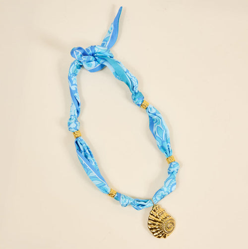 The Darling Effect’s Ocean Muse Make a Statement Scarf Necklace features blue patterned silky fabric, gold beads, and a large gold seashell charm, displayed on a light beige background.