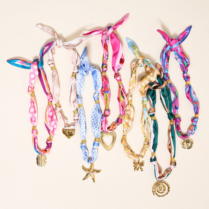 Seven vibrant Ocean Muse Make a Statement Scarf Necklaces by The Darling Effect, featuring silk ribbons, gold bead accents, and assorted gold charms—like hearts, starfish, spirals, and florals—are displayed on a neutral background.