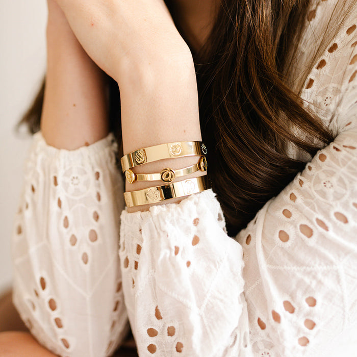 A woman with loose, wavy brown hair rests her chin on her hand, displaying three "Original Trait Cuff Bracelets" by The Darling Effect in gold and sterling silver with engraved designs.