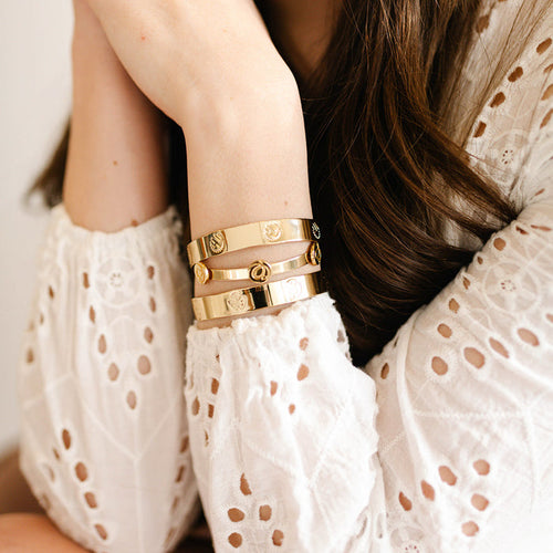 A woman with loose, wavy brown hair rests her chin on her hand, displaying three "Original Trait Cuff Bracelets" by The Darling Effect in gold and sterling silver with engraved designs.
