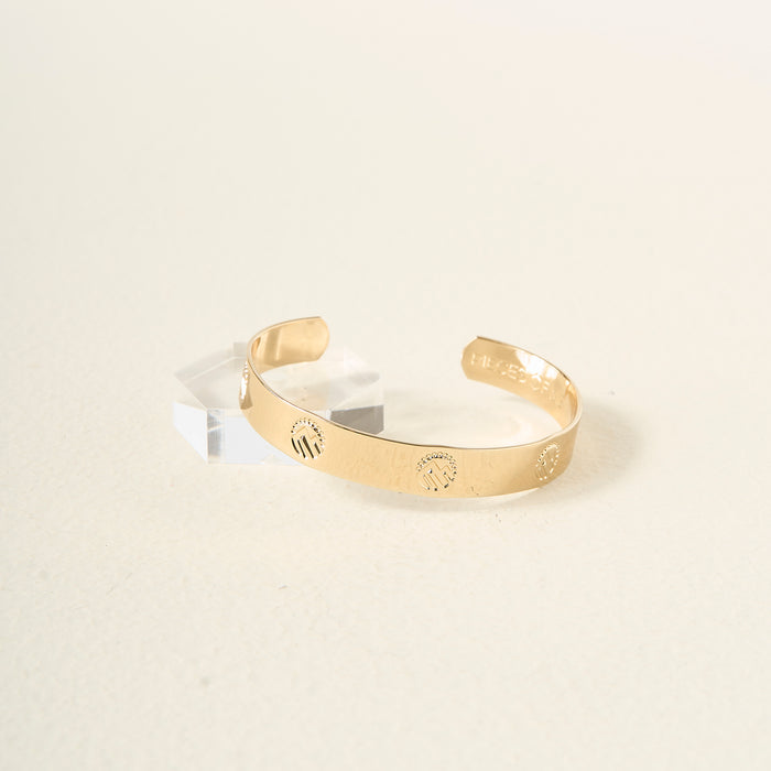 The Darling Effect's Original Trait Cuff Bracelet, featuring engraved designs, is displayed on a light surface and propped against a clear acrylic block, highlighting the elegance of this adjustable accessory.