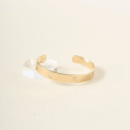 The Darling Effect's Original Trait Cuff Bracelet, featuring engraved markings, is displayed on a light surface beside a clear geometric block, embodying the timeless elegance of this classic accessory.