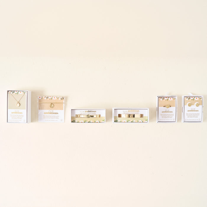 Six jewelry boxes from The Darling Effect are displayed in a row, each holding the Original Trait Cuff Bracelet or other gold-accented floral jewelry such as necklaces and earrings, set against a plain light-colored wall.