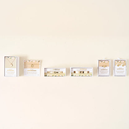 Six jewelry boxes from The Darling Effect are displayed in a row, each holding the Original Trait Cuff Bracelet or other gold-accented floral jewelry such as necklaces and earrings, set against a plain light-colored wall.