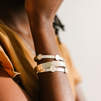 A person in a tan top rests their hand on their shoulder, showcasing three metallic bracelets—including The Darling Effect’s Original Trait Cuff Bracelet—on their wrist. The softly lit background is neutral.