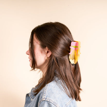 A woman with brown hair, viewed from the side, wears The Darling Effect’s Pencil Me In Shape Up Claw Clip—a yellow pencil-shaped accessory with a pink eraser—paired with a light blue denim jacket against a plain beige background.