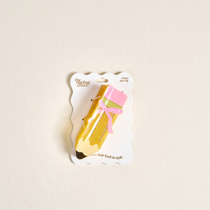 Add a playful touch to your look with The Darling Effect's Pencil Me In Shape Up Claw Clip, featuring a cute pencil design with pink eraser and bow—charmingly displayed on white wavy-edged packaging against a light beige background.