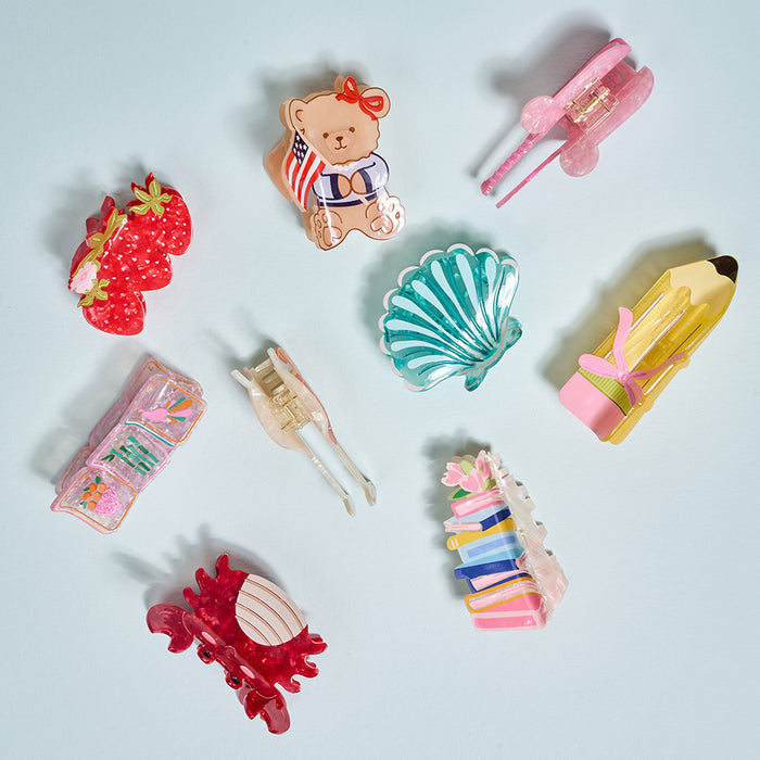 The Darling Effect’s Pencil Me In Shape Up Claw Clip is featured among colorful, cute hair clips in playful shapes like teddy bears, strawberries, shells, pencils, books, and abstract designs on a light blue background.