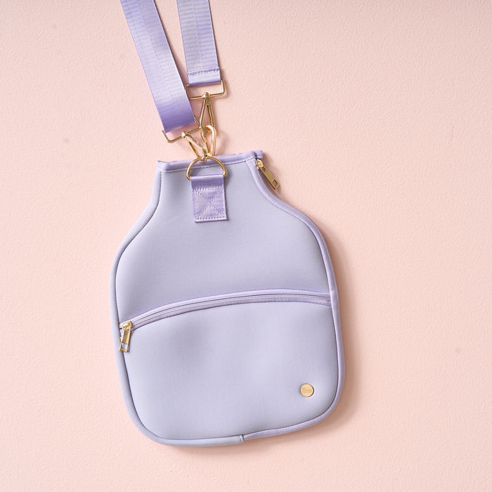 The Darling Effect’s Periwinkle Pickleball Paddle Case, a light purple crossbody with gold zipper and round button, hangs from a matching adjustable strap against a pale pink background.