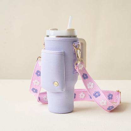 The Darling Effect’s Periwinkle Tumbler Sling holds a lavender tumbler with white lid and straw in a matching neoprene sleeve with side pocket, an adjustable pink strap with purple and white flowers, gold clasps, and an off-white background.