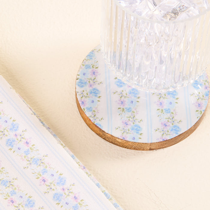 A ribbed glass filled with ice rests on the-darling-effect’s Petal Parade Blue Wood Enamel Coaster. Matching blue floral-patterned fabric or paper appears partially to the left on a light cream surface.