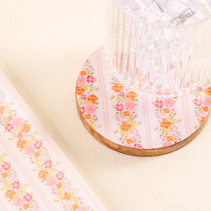 A glass of ice water rests on the Petal Parade Pink Wood Enamel Coaster by the-darling-effect, featuring pink stripes and orange florals—proof that decorative coasters can brighten any setting.