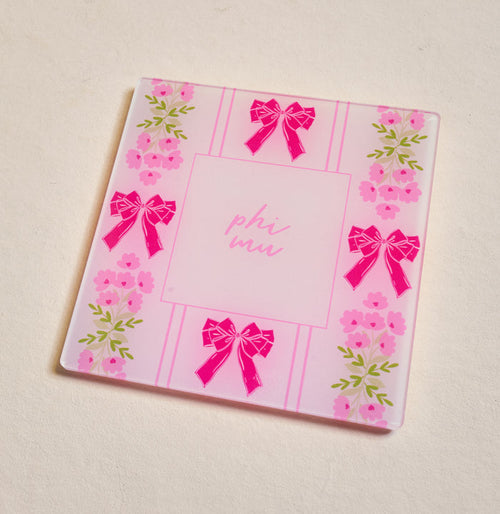 The Darling Effect's Phi Mu Acrylic Coaster is a square pink coaster featuring "phi mu" in the center with bows and floral edges—ideal for chapter pride or as a cute Big/Little gift, beautifully displayed on a light surface.