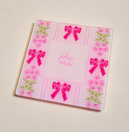 The Darling Effect's Phi Mu Acrylic Coaster is a square pink coaster featuring "phi mu" in the center with bows and floral edges—ideal for chapter pride or as a cute Big/Little gift, beautifully displayed on a light surface.