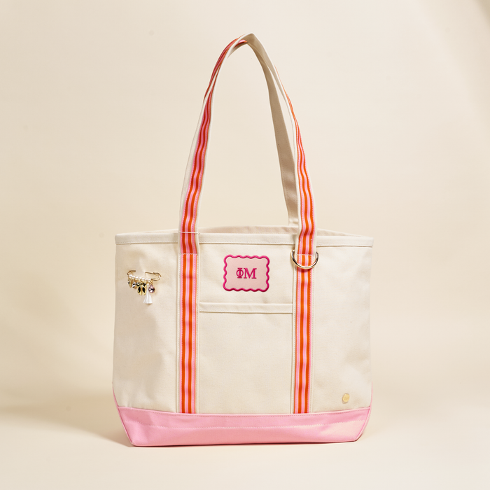 The Darling Effect's Phi Mu Canvas Tote Bag is a beige canvas tote with pink accents and striped handles. It showcases a monogrammed M in a pink frame, plus a gold charm on one handle—ideal as a stylish sorority bag.