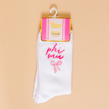 The Darling Effect Phi Mu Embroidered Crew Socks feature white fabric, pink bow detail, and come in pink-yellow packaging labeled "that’s so sorority socks," making them a perfect sorority gift. They hang on a white plastic hook.